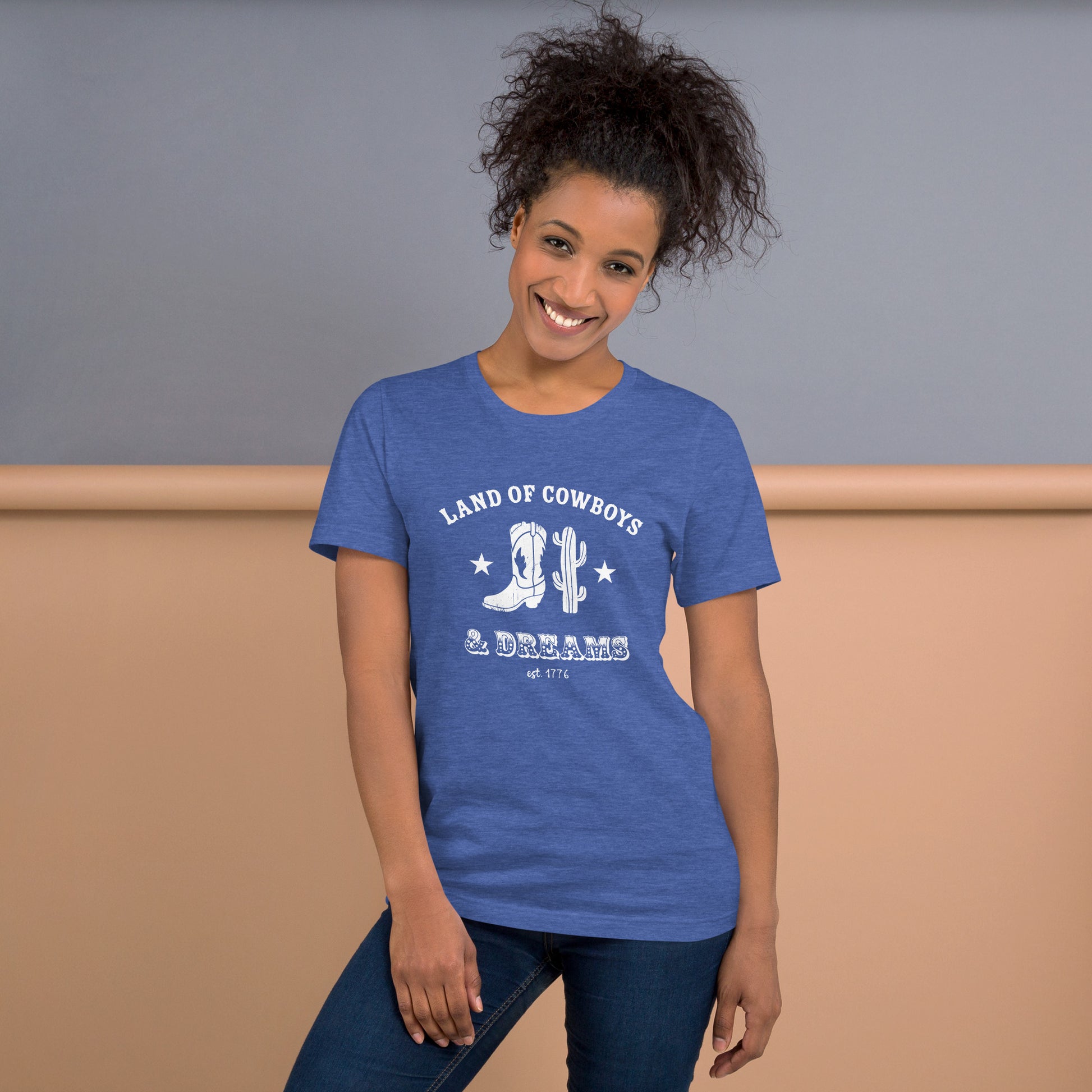 Woman wearing a blue t-shirt with a white graphic featuring a cowboy boot, cactus, and text that says “Land of the Cowboys and Dreams est. 1776.”