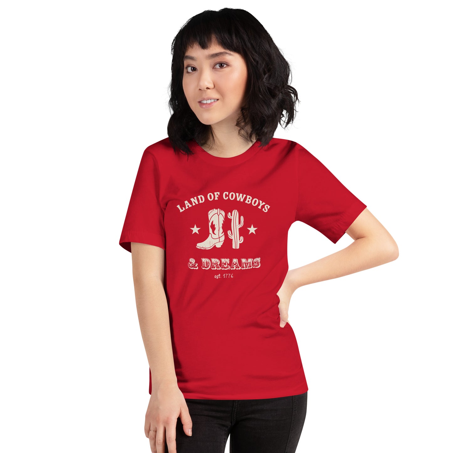Woman wearing a red t-shirt with a white graphic featuring a cowboy boot, cactus, and text that says “Land of the Cowboys and Dreams est. 1776.”