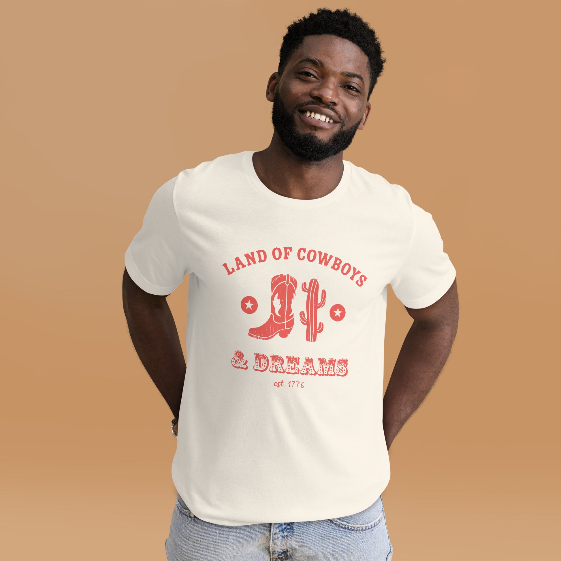 Man wearing a white t-shirt with a red graphic featuring a cowboy boot, cactus, and text that says “Land of the Cowboys and Dreams est. 1776.”