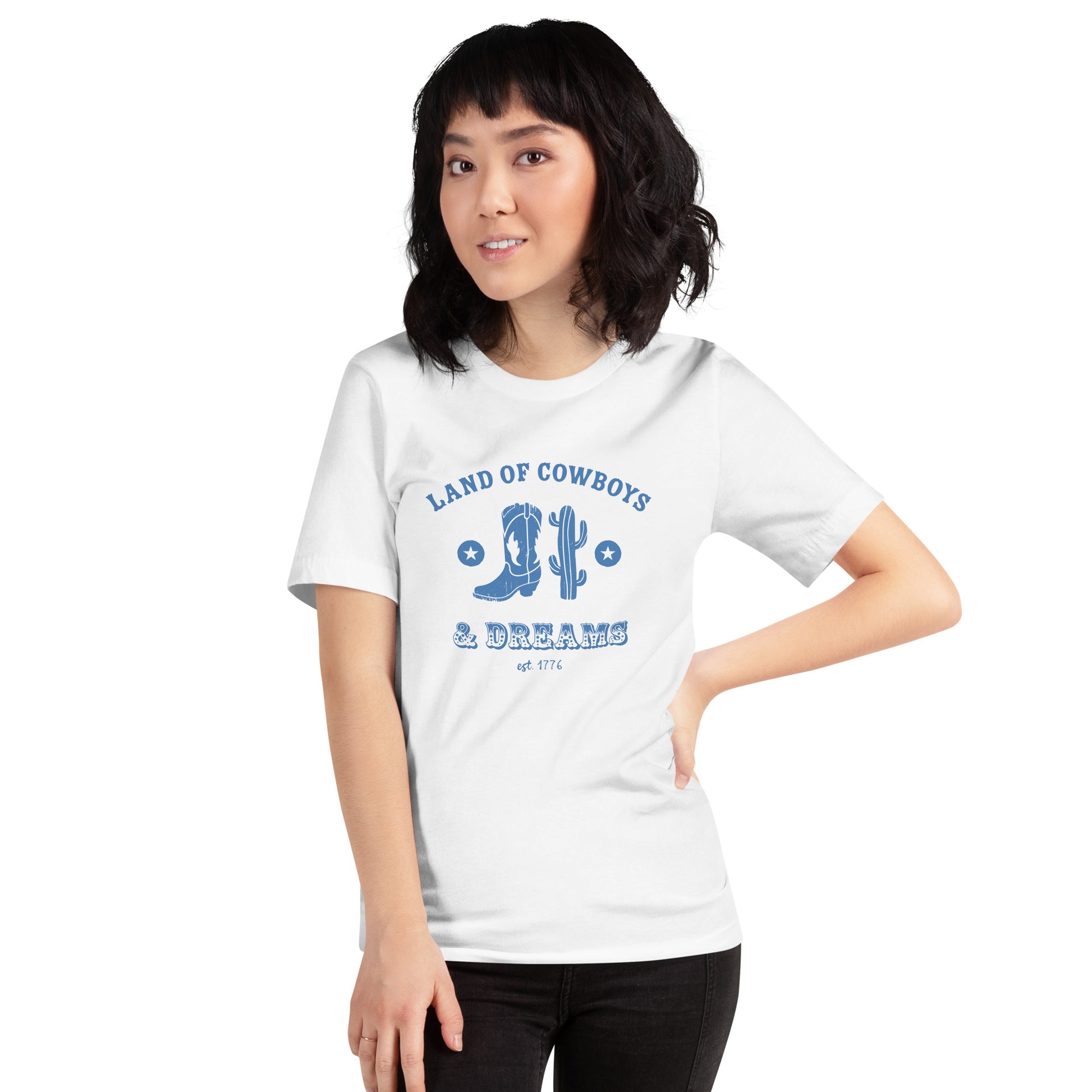 Woman wearing a white t-shirt with a blue graphic featuring a cowboy boot, cactus, and text that says “Land of the Cowboys and Dreams est. 1776.”