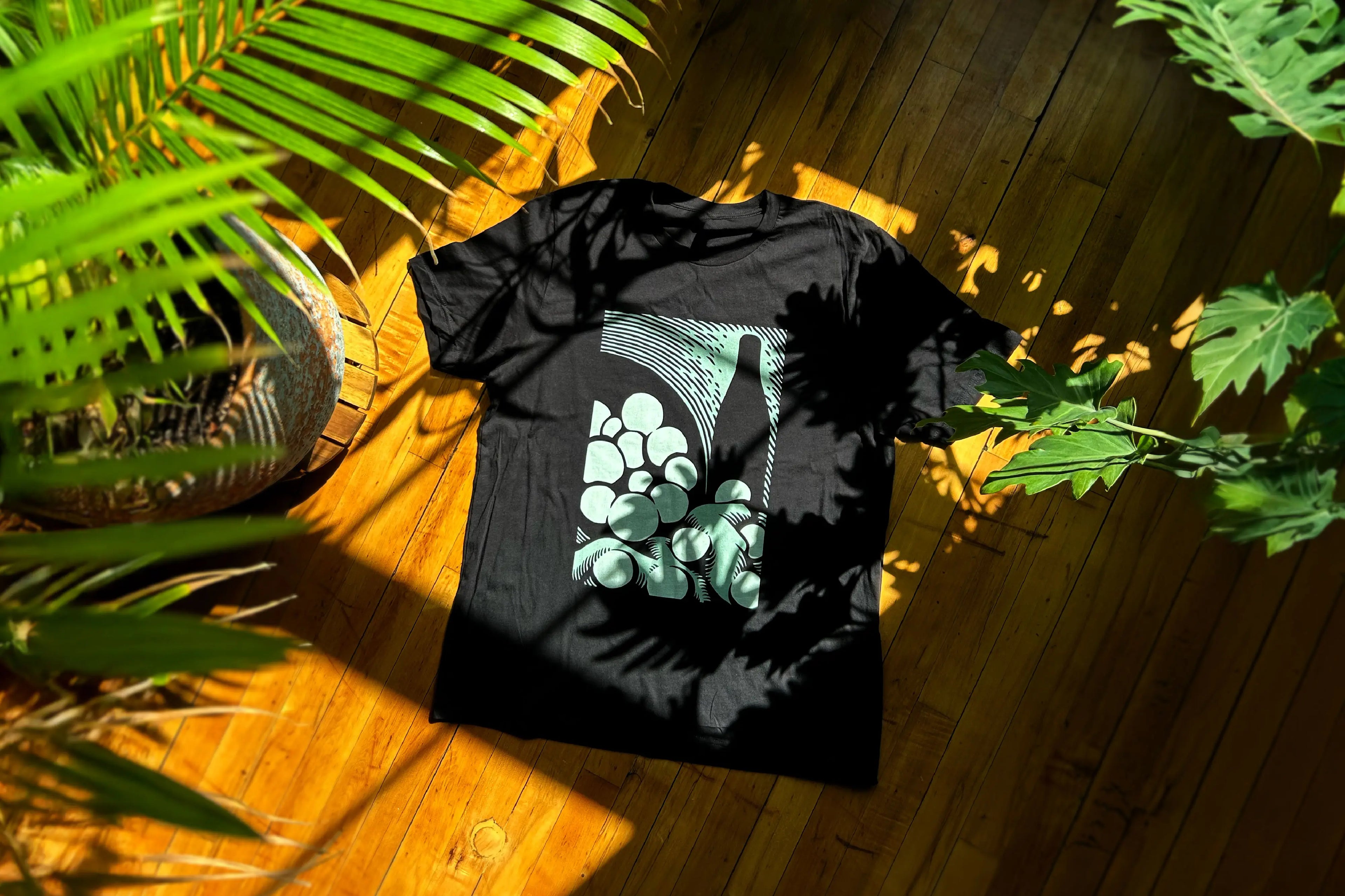 Black t-shirt with green graphic design on wooden floor with leaf shadows
