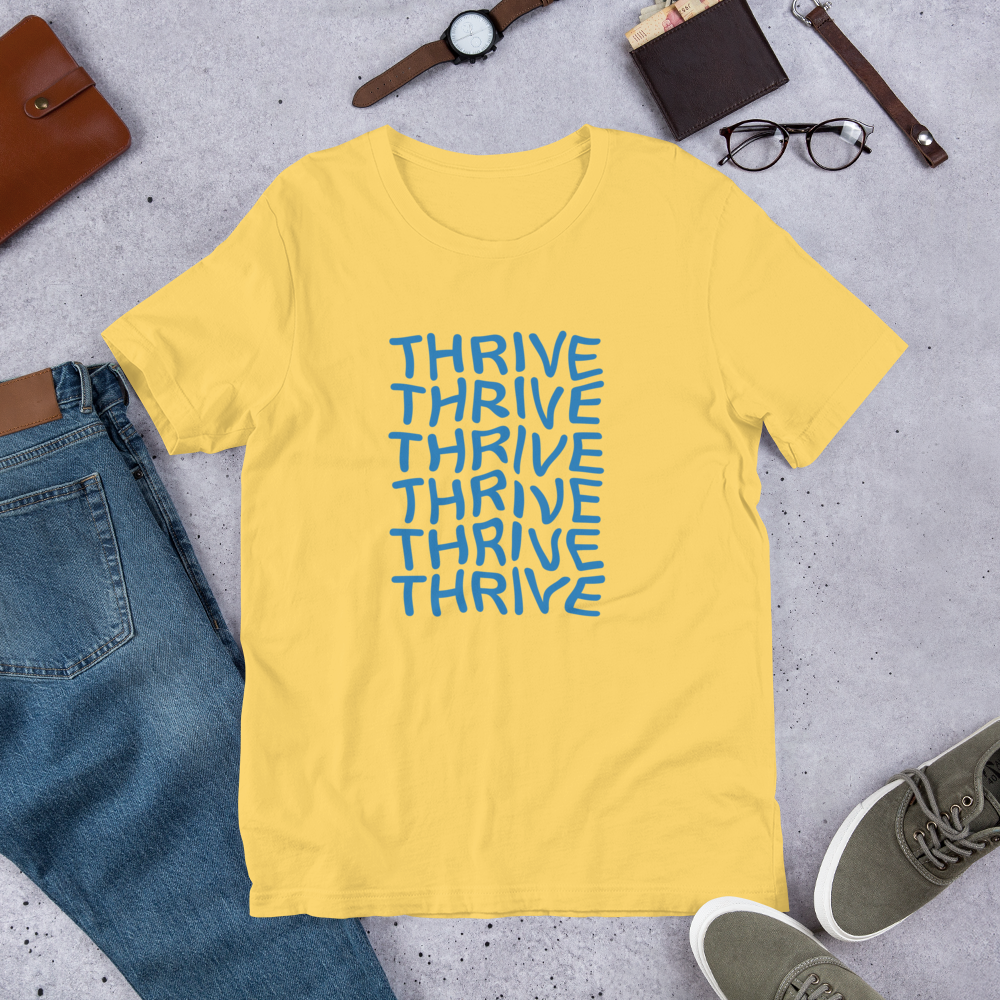 Yellow t-shirt laid flat next to a pair of jeans and accessories. The shirt design features the word 'Thrive' written six times stacked on top of each other in white text.